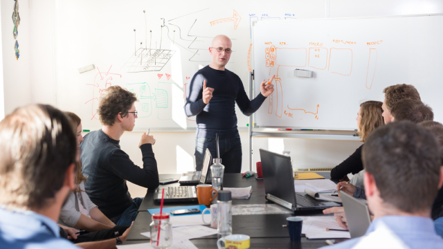 An instructor stands at a large paperboard with various notes, speaking to a class of learners seated on either side of a large table