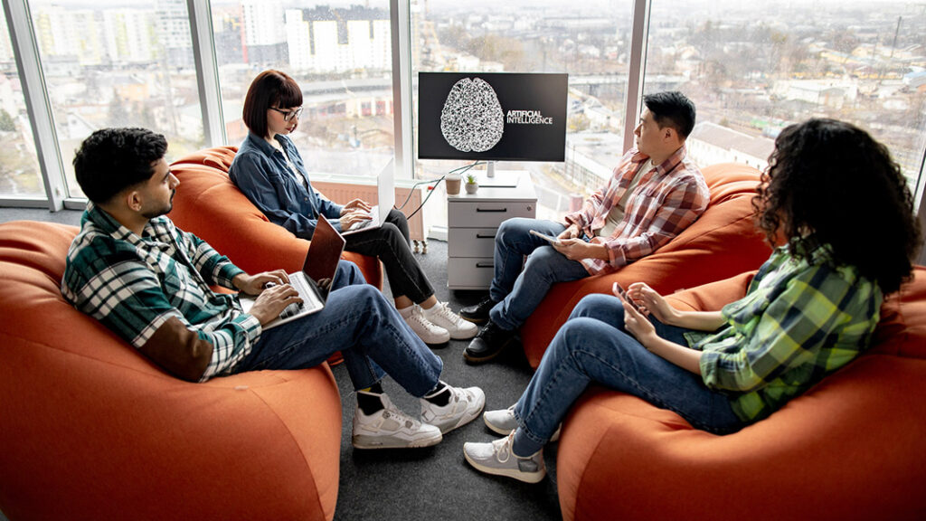 People sitting on bean bags working on their tech devices - windows in background showing city scape and a large screen displaying a vector of a brain with Artificial Intelligence as the title