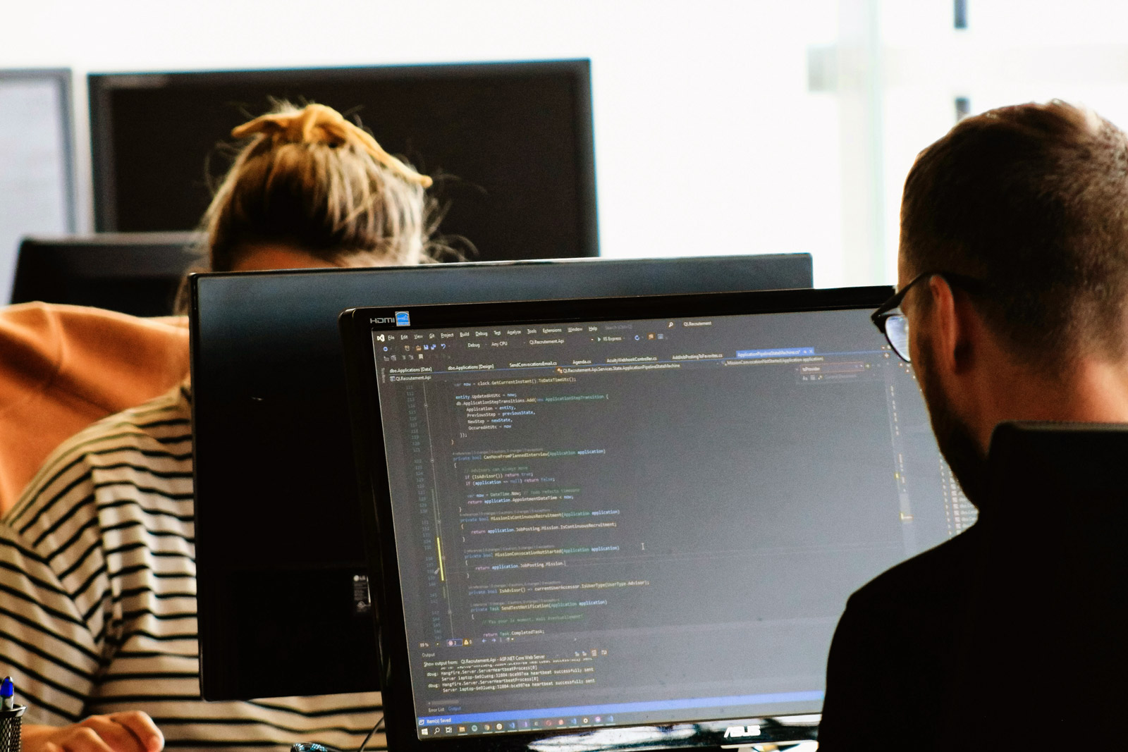 man sitting at a computer with his back us, woman seen in background at a computer
